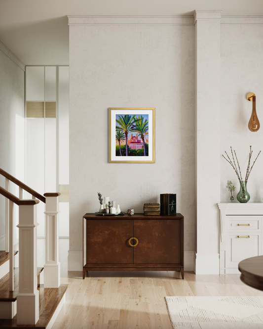Staircase with a wooden sideboard and decorative items in a home interior. A gold framed artwork print with palm trees and an ornate pink building.