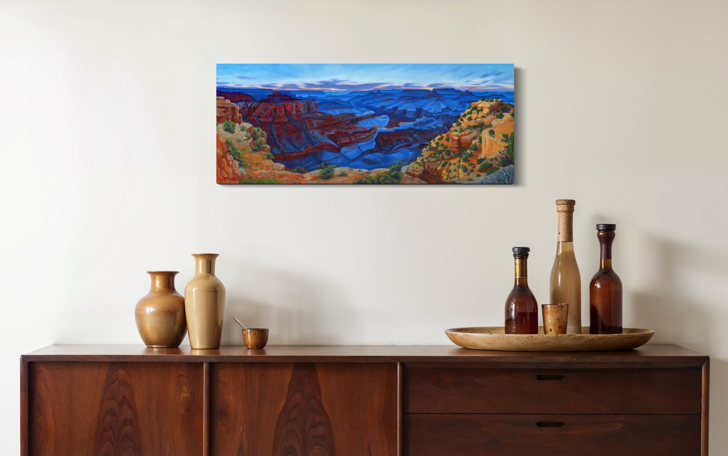 Painting of a landscape above a wooden sideboard with decorative items. Painting is of the Grand Canyon with red and orange cliffs and blue shadows just after sunset.