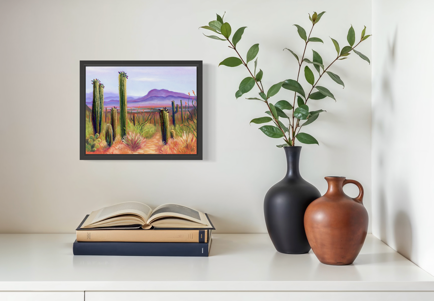 Framed artwork of a desert landscape on a shelf with books and vases.