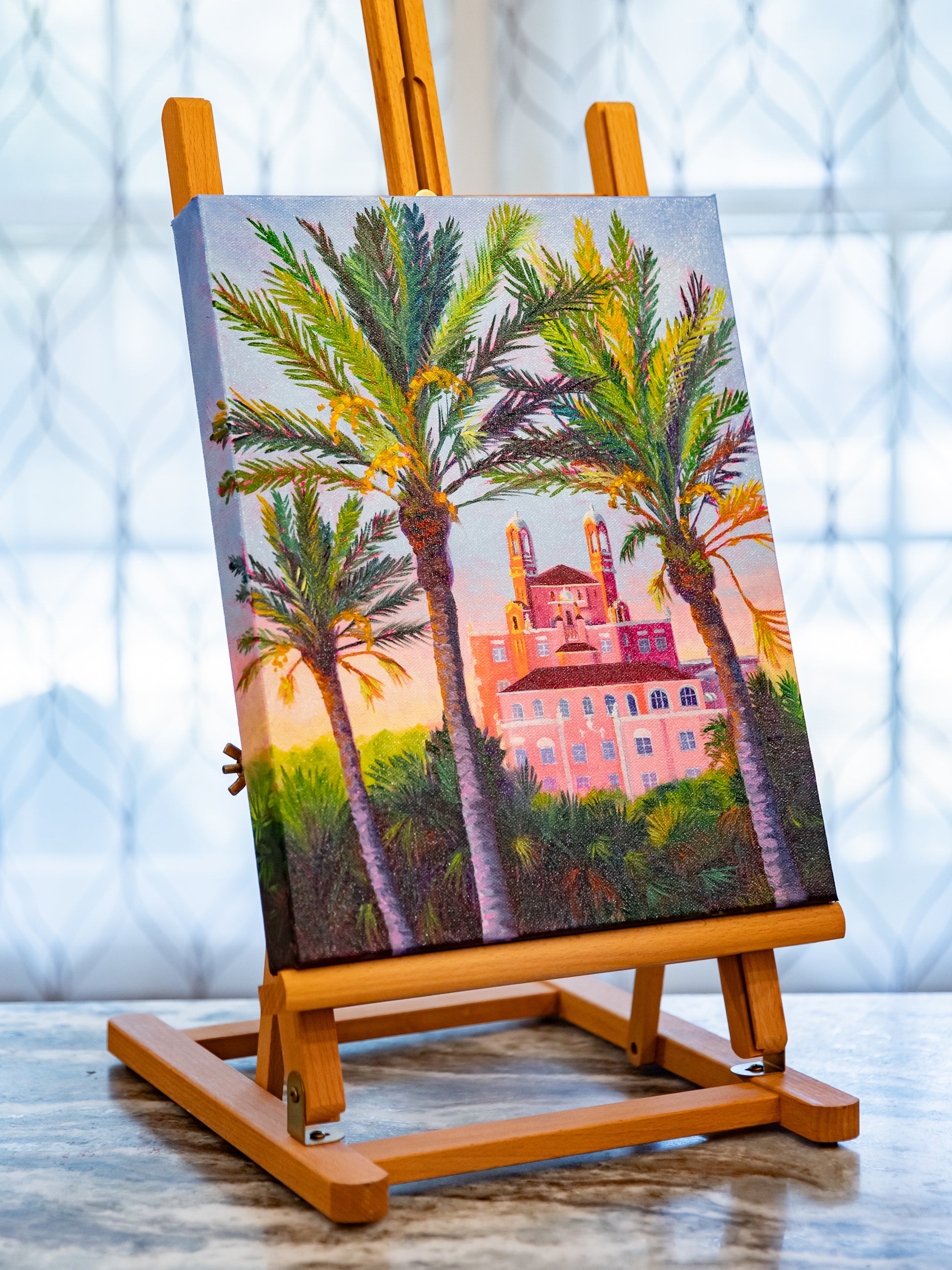 Painting of palm trees and a pink building on an easel with a blurred indoor background
