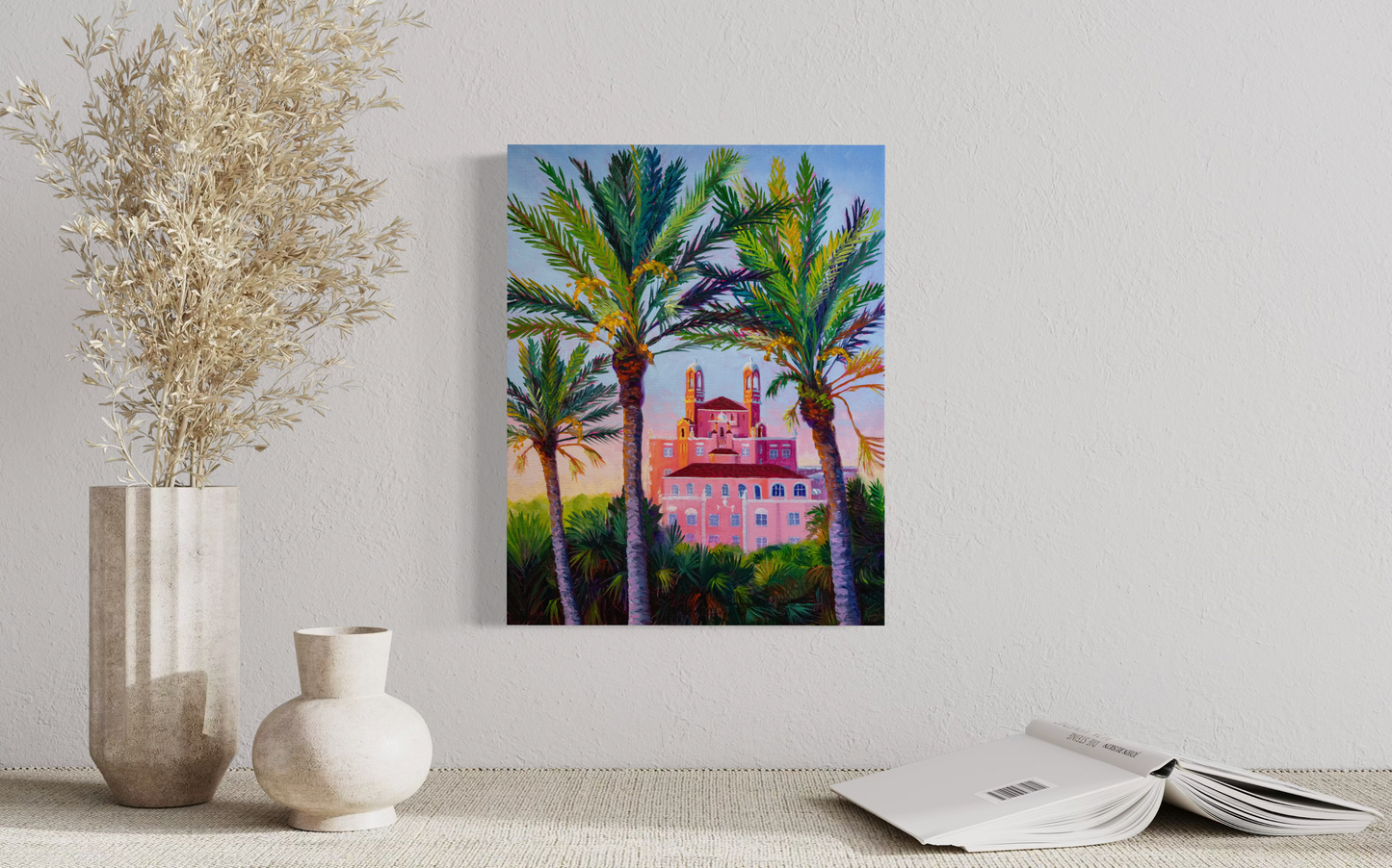 Painting of palm trees and a pink ornate building on a wall above two vases and an open book.