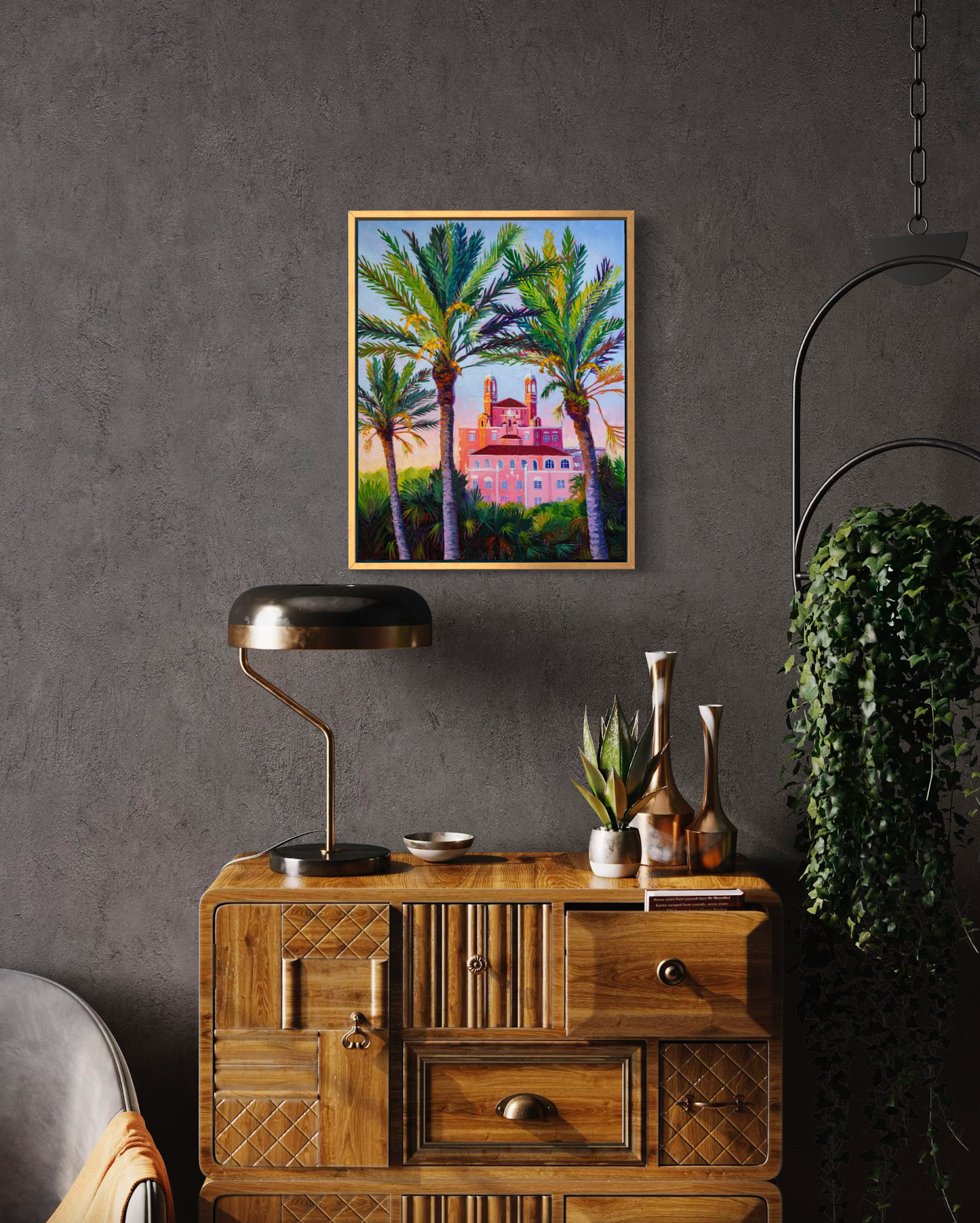 Decorative interior with wooden sideboard, lamp, and painting of palm trees and a building on a gray wall.