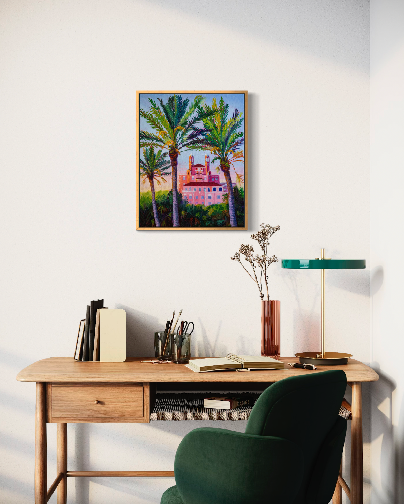 Home office with a desk, chair, and wall art of palm trees and a building printed on canvas framed in gold.