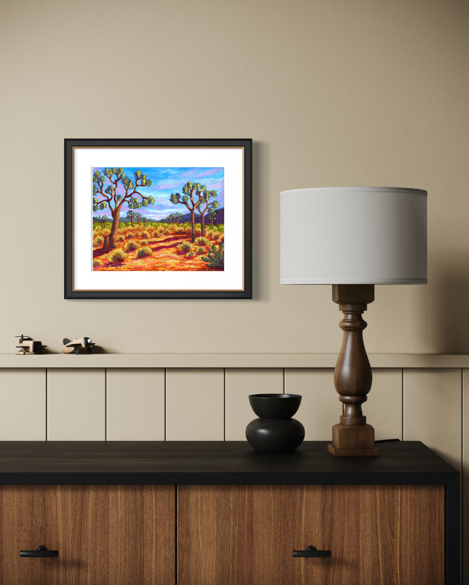 Framed artwork of a desert landscape on a wall above a wooden cabinet with a lamp.