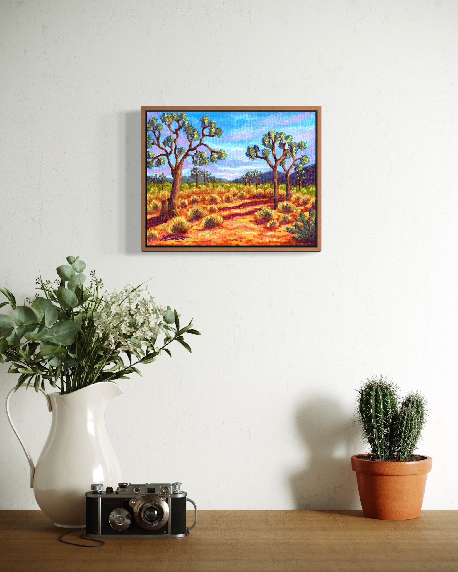 Framed desert landscape painting on a wall with a pitcher, camera, and cactus below.