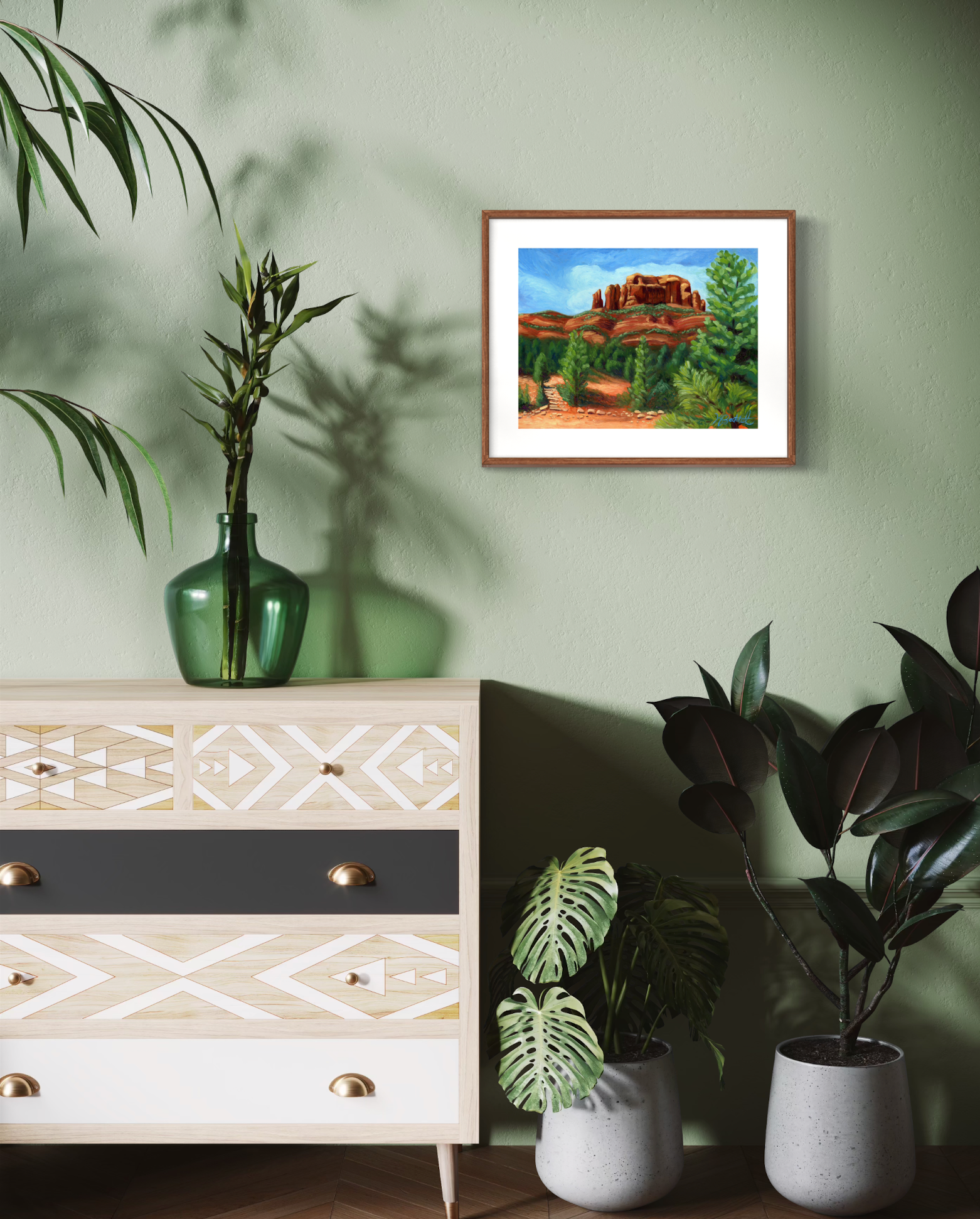 Framed artwork of a natural landscape on a green wall above a dresser with decorative plants.