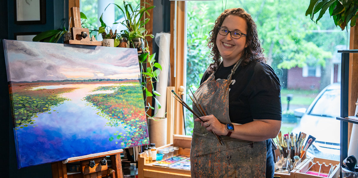 A woman wearing glasses, a black t-shirt, and a brown paint-splattered apron stands in an artist studio holding paintbrushes next to a colorful landscape painting on an easel.