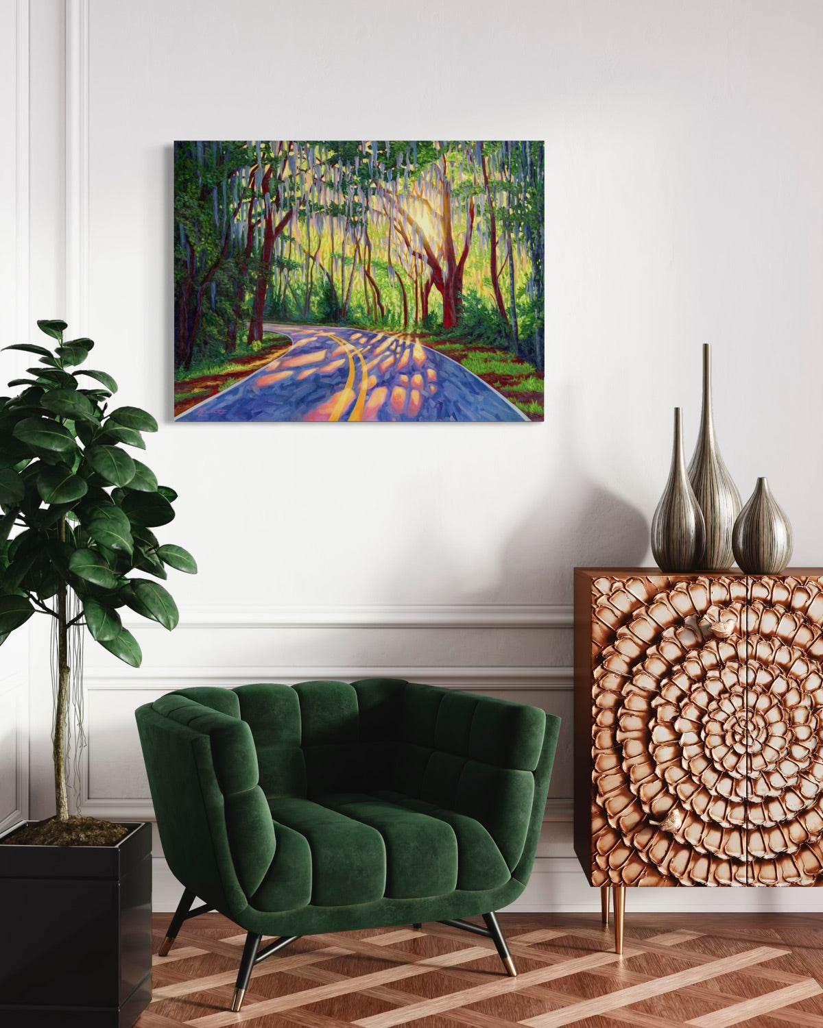 A colorful canvas painting of a sunlit forest road with trees and blue-purple shadows, displayed on a white wall above a green velvet armchair in a styled room with a wood cabinet and plant.