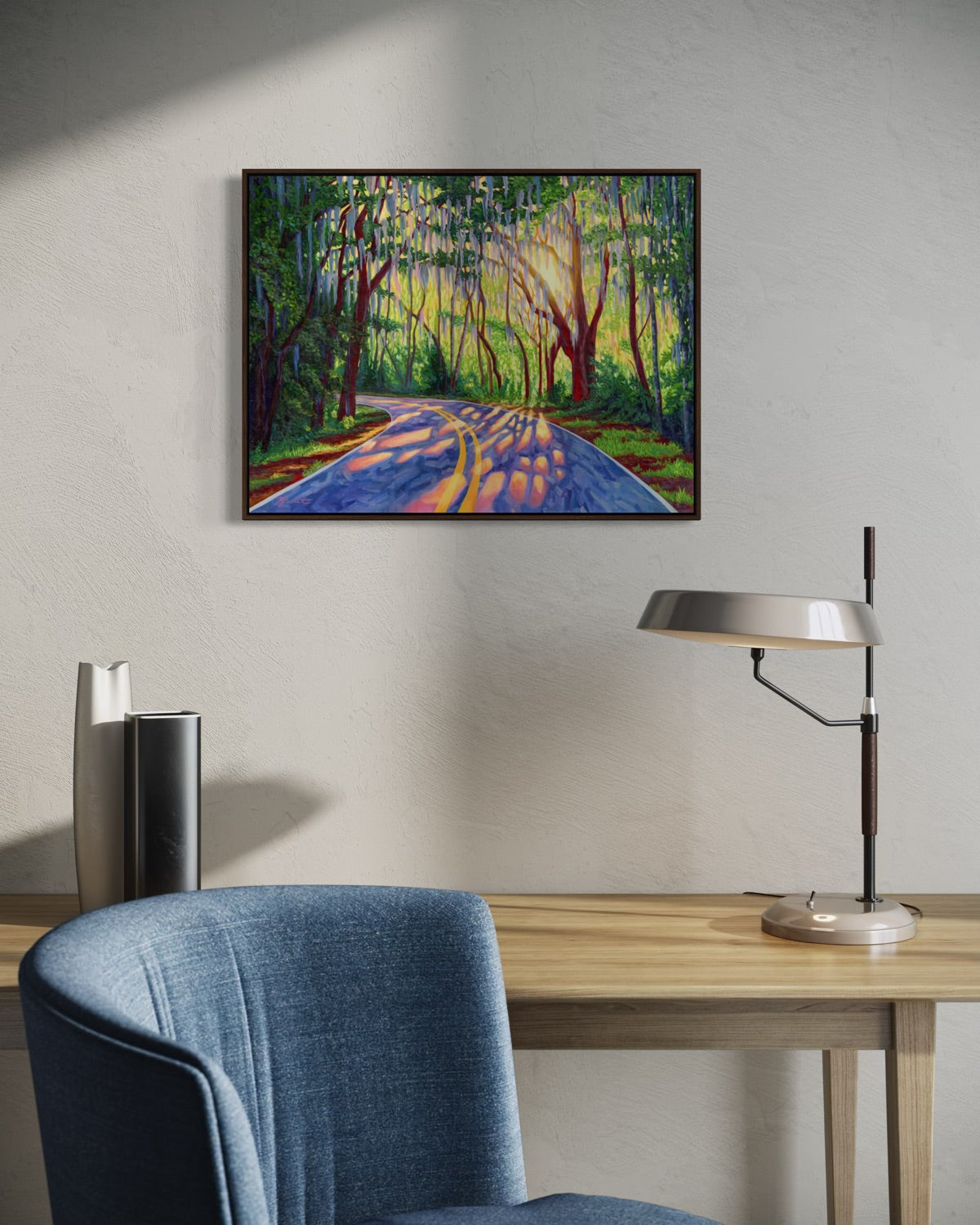 A framed colorful landscape painting of a sunlit road winding through a forest with hanging mossy trees, displayed on a light gray wall above a wooden desk with a blue chair and a silver desk lamp.