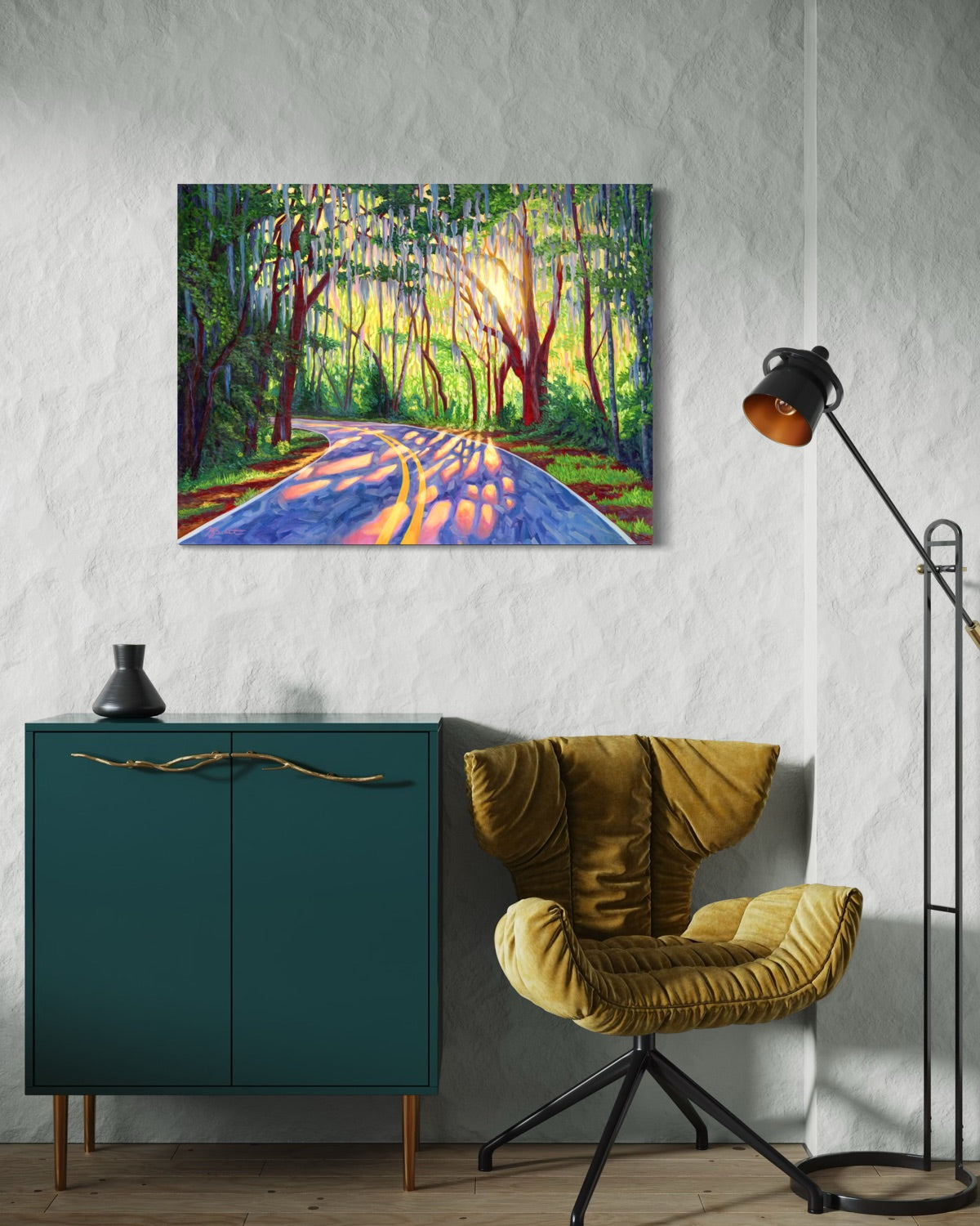 A colorful  painting depicting a sunlit forest road with hanging mossy trees, displayed above a teal cabinet in a styled room with a mustard velvet chair and a black floor lamp.