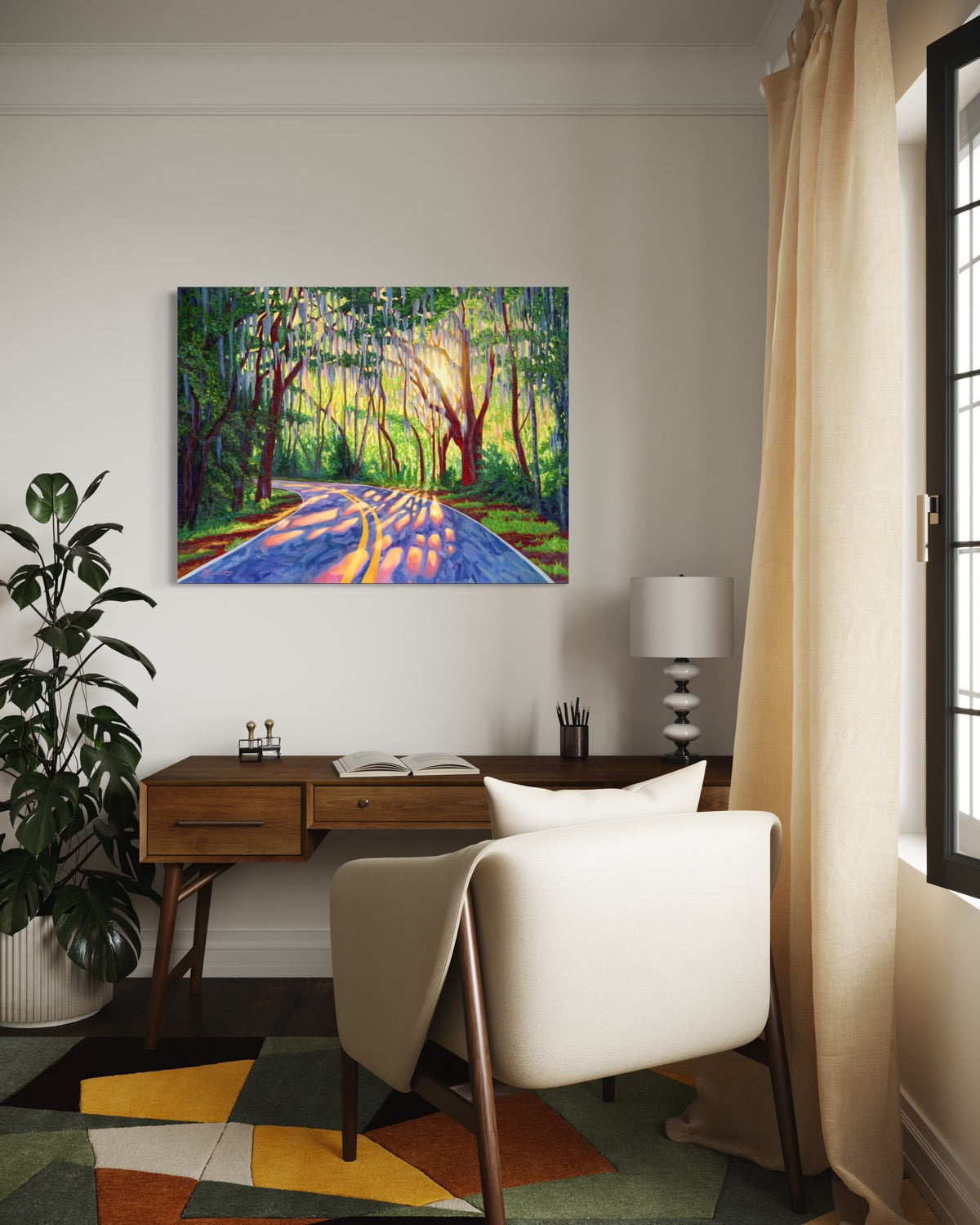 A colorful landscape canvas painting of a sunlit forest road with Spanish moss trees, displayed on a white wall in a modern home office setting above a wooden desk and cream chair.