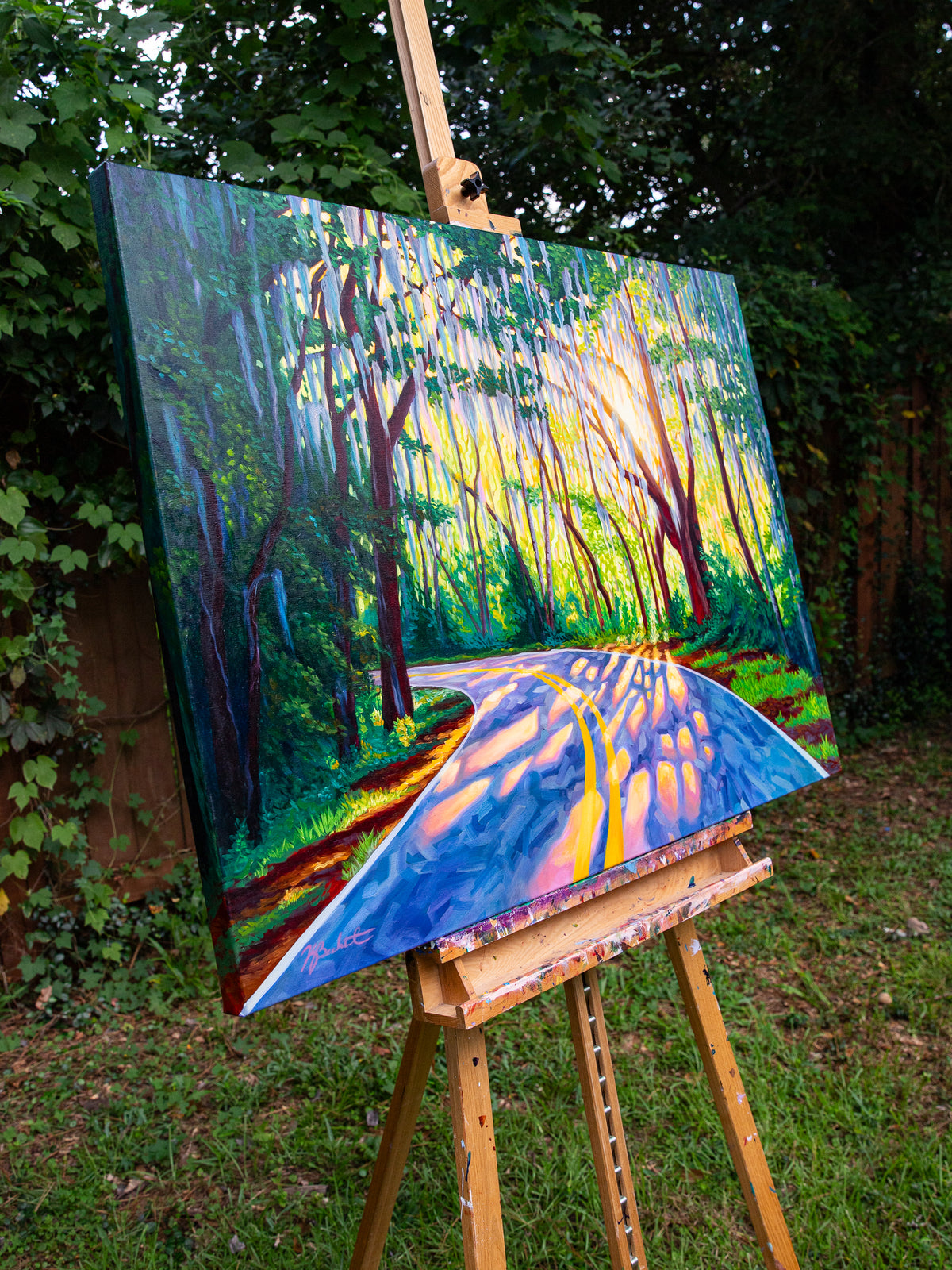 A colorful landscape painting of a sunlit forest road with trees and vivid blue, yellow, green, and orange tones, displayed on a wooden easel outdoors in a garden setting.