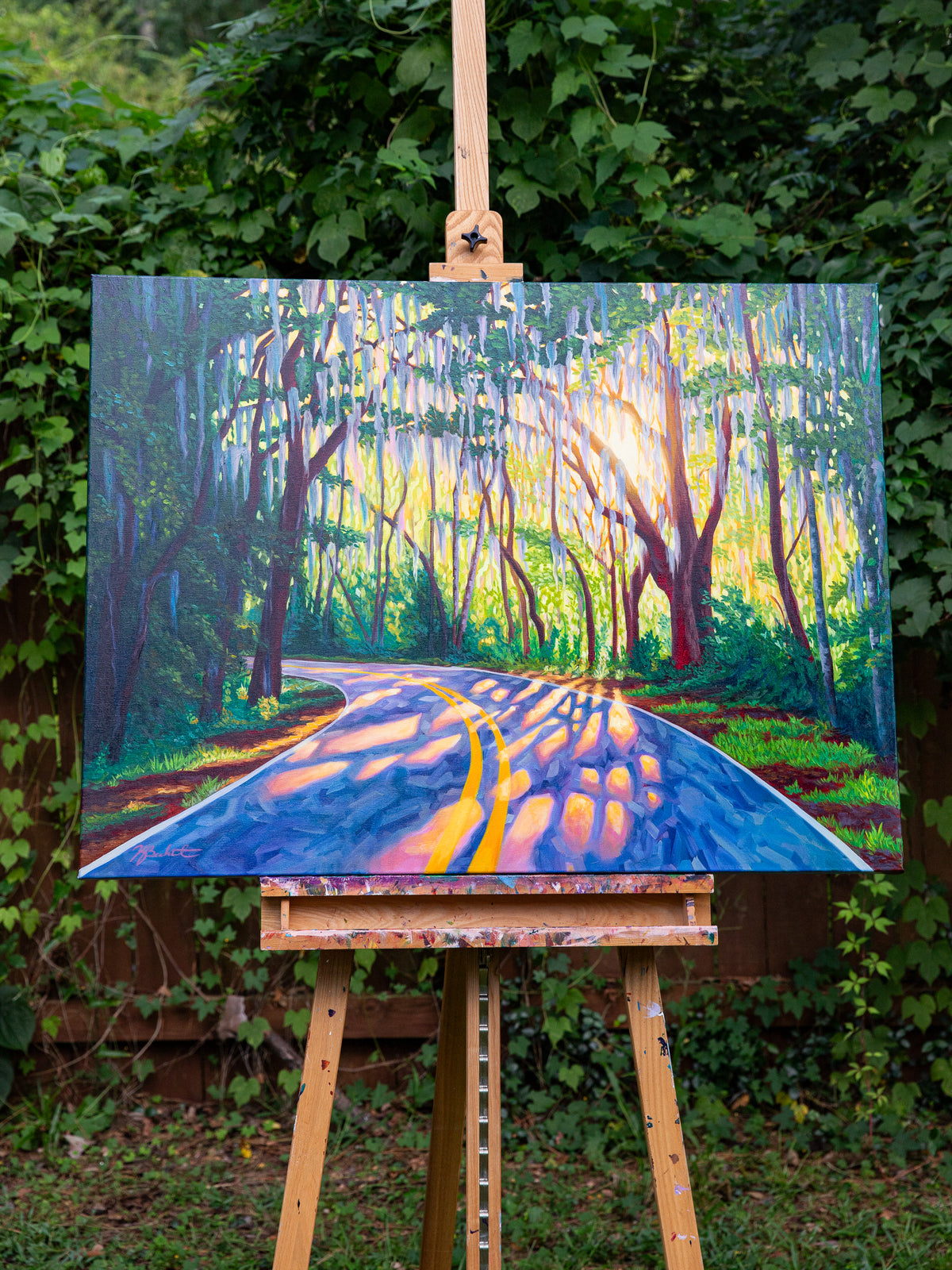 A colorful painting of a winding road through a forest with hanging cypress trees and bright sunlight, displayed on a wooden easel outdoors in front of green foliage.