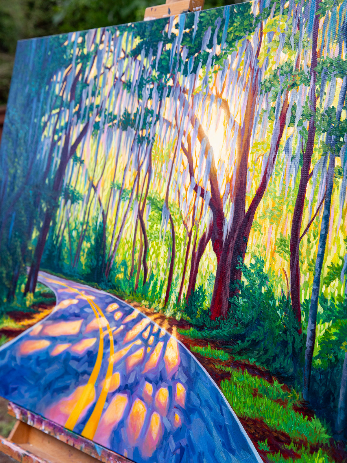 A colorful landscape painting of a sunlit forest road with tall trees, shown on an easel. The artwork features vivid greens, yellows, blues, and purples, with bright sunlight filtering through the trees.