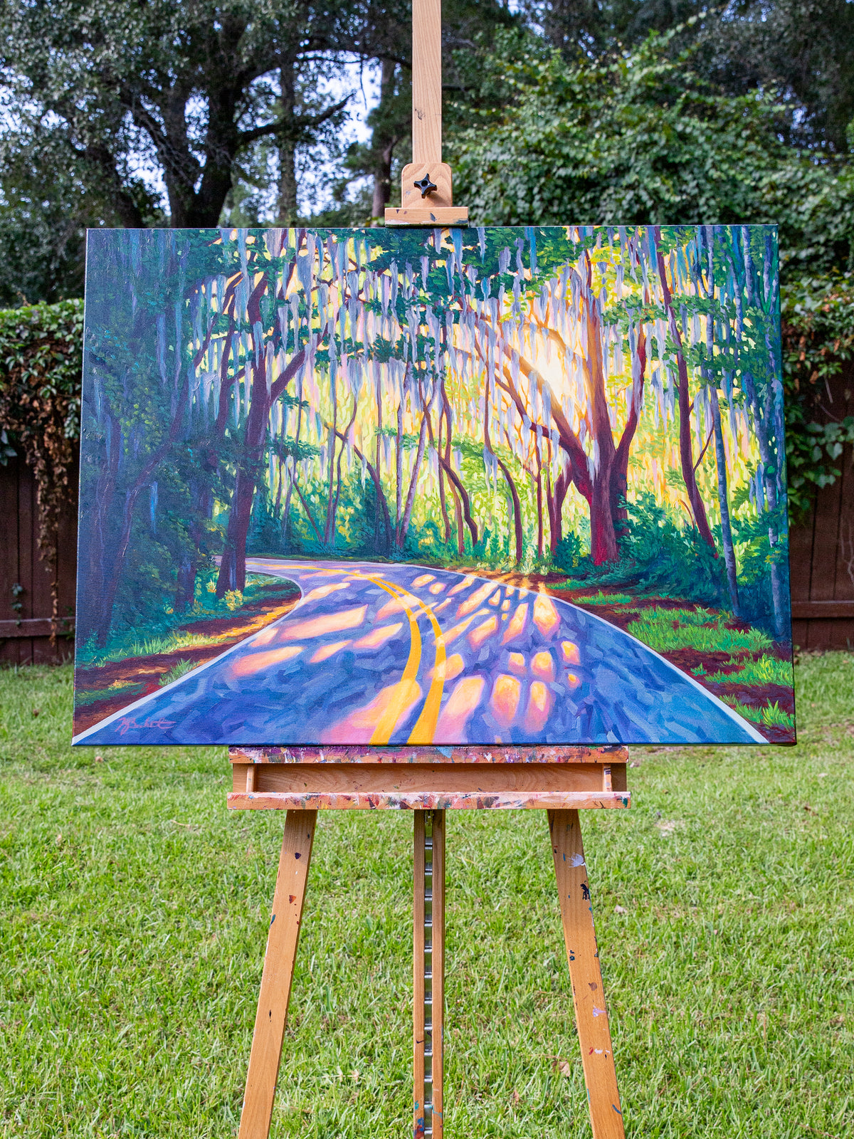 A colorful landscape painting of a sunlit road winding through a forest, displayed on a wooden easel outdoors on a grassy lawn.