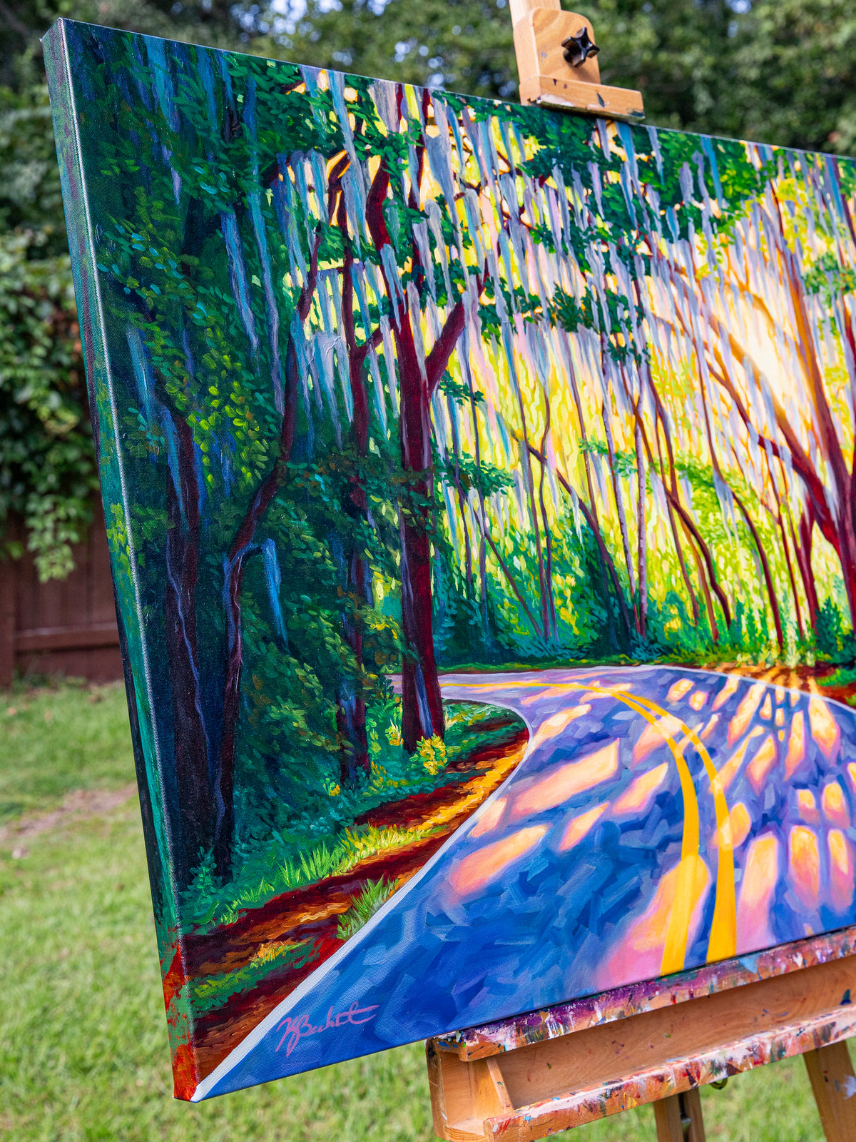 A colorful landscape painting of a winding road through a forest, displayed on an artist's easel outdoors with grass and trees in the background.