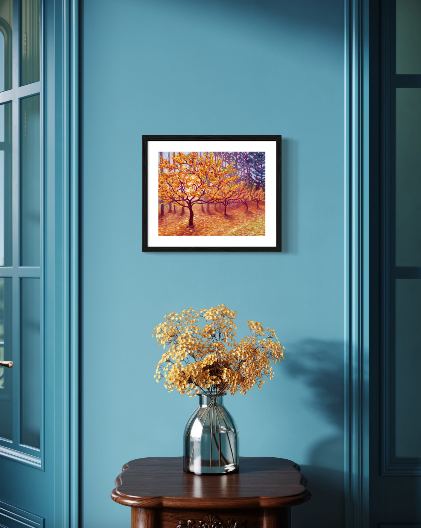 Framed artwork of a tree with autumn leaves on a blue wall above a vase with flowers.