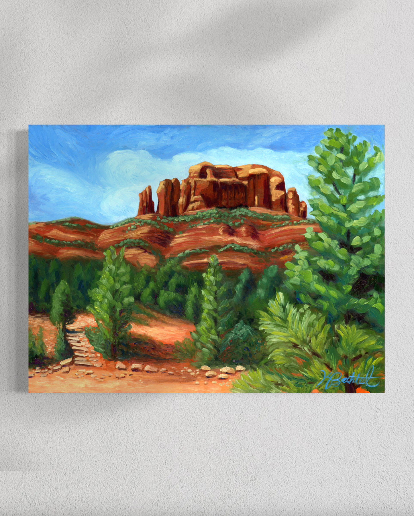 Painting of a desert landscape with red rock formations and green trees on a white wall.