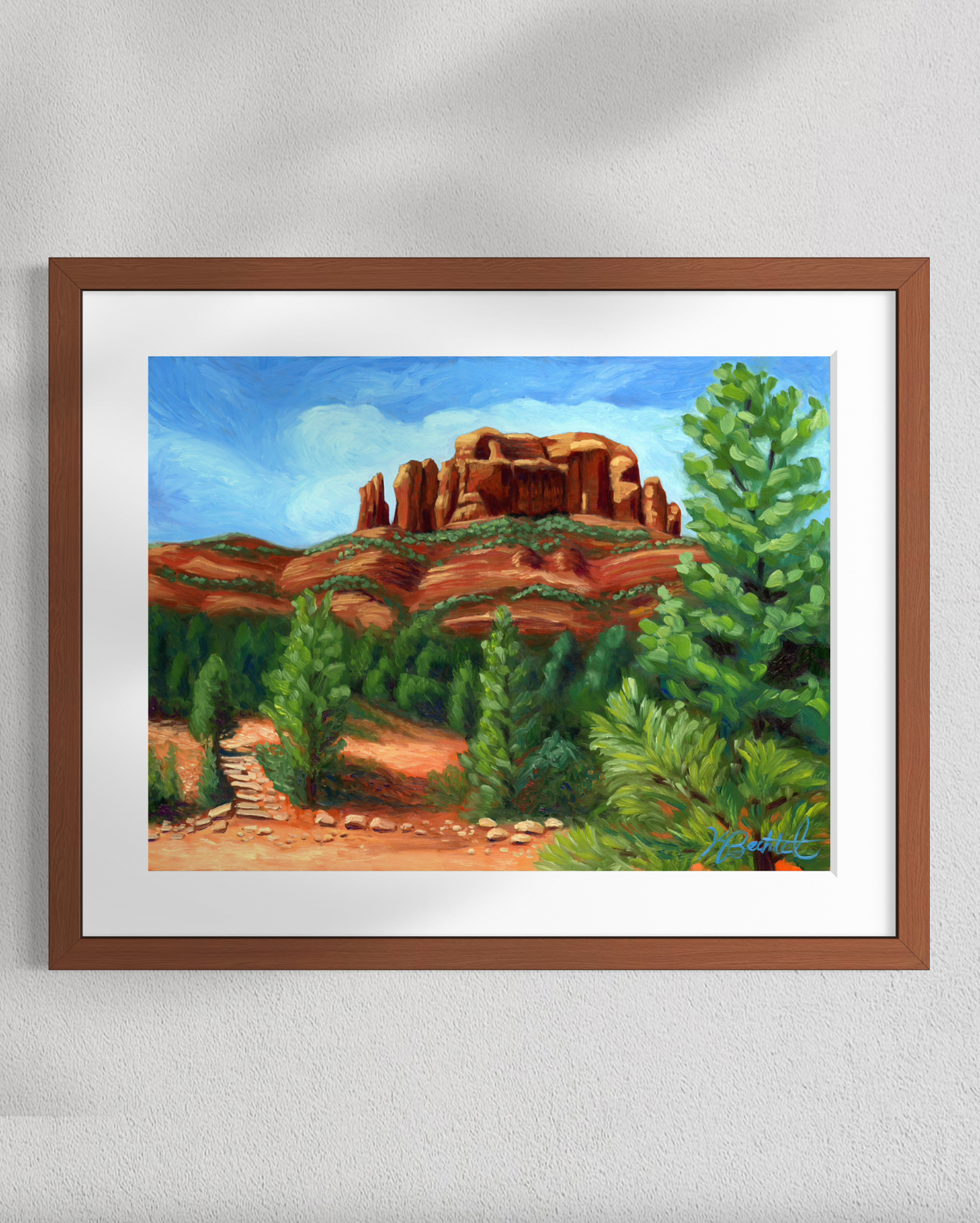 Framed painting of a desert landscape with red rock formations and green trees on a white wall.